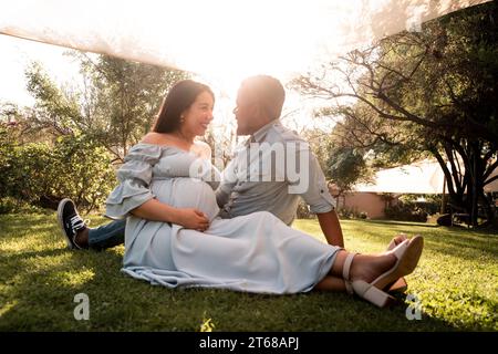 Eine schwangere Latina-Frau und ein afrikanisches Männerpaar sitzen in einem üppig grünen, hellen, sonnendurchfluteten Park Stockfoto