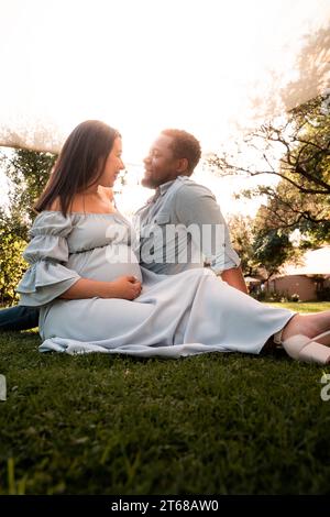 Eine schwangere Latina-Frau und ein afrikanisches Männerpaar sitzen in einem üppig grünen, hellen, sonnendurchfluteten Park Stockfoto