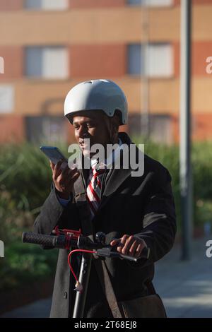 Ein junger Mann in einer professionellen Anzugsjacke und einem weißen Helm, der auf einem Roller steht und sein Handy benutzt Stockfoto
