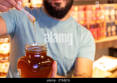 Der Mann hält ein Glas Honig und zeigt, wie der Honig tropft, während er einen Löffel davon hebt, vor dem Hintergrund eines Ladenregals Stockfoto