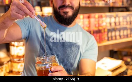 Der Mann hält ein Glas Honig und zeigt, wie der Honig tropft, während er einen Löffel davon hebt, vor dem Hintergrund eines Ladenregals Stockfoto