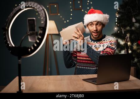 Glücklicher arabischer männlicher Blogger, der Geschenkboxen von Firmen auspackt und Livestreaming macht. Influencer mit Smartphone-Kamera zum Erstellen von Inhalten Stockfoto