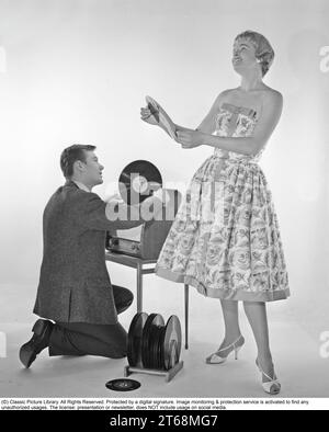 Grammophon in den 1950er Jahren Ein Mann kniet bei einem kombinierten Repetierset und einem Grammophonschrank, wo er die Abdeckung des Grammophons geöffnet hat und eine Platte in der Hand hält. Sie trägt ein typisches Kleid aus den 1950er Jahren und sieht glücklich aus. Schweden 1959. Kristoffersson Ref. CD65-3 Stockfoto
