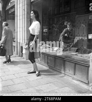 Junge Frau 1956. Sie steht vor einem Schaufenster in Strandvägen in Stockholm und vielleicht weiß sie nicht, wie der Typ im Teppichladen im Fenster sitzt und sie bewundernd ansieht. Sie trägt einen weißen Pullover und eine kniehohe Hose. Schweden 1956. Kristoffersson Ref. BS94-3 Stockfoto