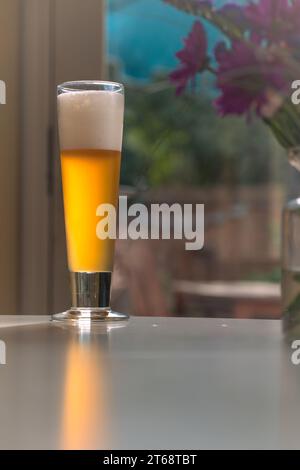 Ein klares Bierglas mit weißem Schaumkopf steht auf einem Holztisch vor einer Fensterbank mit einem bunten Blumenstrauß Stockfoto