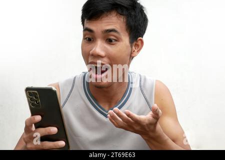 Nahaufnahme Porträt eines jungen asiatischen Mannes isoliert auf weißem Hintergrund, der schockiert aussieht, während er sein Smartphone lächelnd und lachend ansieht Stockfoto