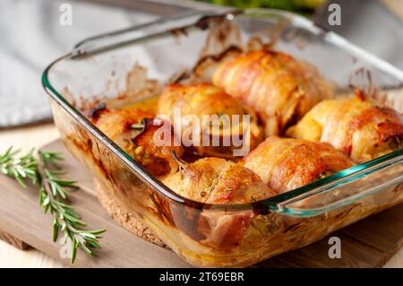 Mit Speck umwickelte Hähnchenschenkel ohne Haut mit geröstetem Brokkoli und Kräutern in einer quadratischen Glasbackform Stockfoto