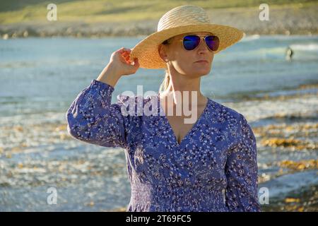 Eine Frau, die in einem blauen Kleid am Strand steht und einen Strohhut mit der Hand hält. Stockfoto