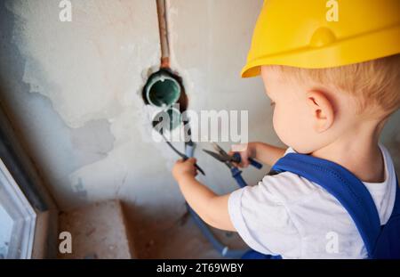 Nahaufnahme eines Babyelektrikers, der Drähte mit einer Zange im Bau schneidet. Kind in einem Schutzhelm, der Kabel in der Wand befestigt, während es an der Renovierung zu Hause arbeitet. Stockfoto