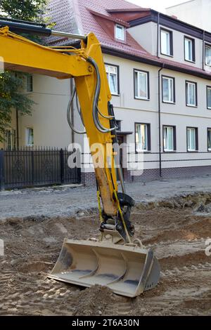 Der Eimer eines Baggers, der mit der Reparatur und dem Verlegen einer Straße vor dem Hintergrund von Häusern auf einer Stadtstraße beschäftigt ist. Stockfoto