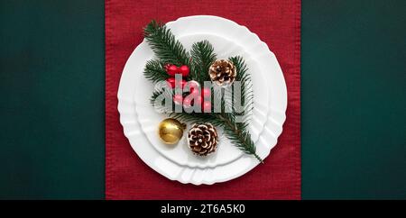Weihnachtstisch mit Dekoration, weißer leerer Teller auf roter Tischdecke, grüner Hintergrund, Draufsicht, oben. Weihnachtsfeier: Arrangement, rot A Stockfoto