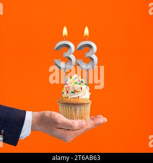 Die Hand, die Cupcake mit der Zahl 33 Kerze liefert - Geburtstag auf orangem Hintergrund Stockfoto