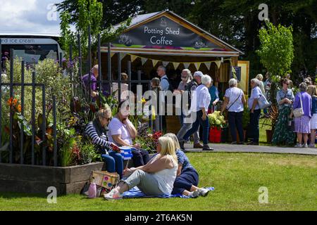 Besucher kommen vorbei an einem Kaffeestand und vier Picknickern, die auf dem Rasen auf dem geschäftigen, sonnigen Ausstellungsgelände sitzen - 2023 Flower Show Tatton Park, Cheshire England, Großbritannien. Stockfoto