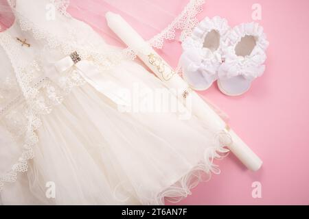 Baby-Taufkleid auf rosa Hintergrund mit Kreuz, Kerze und Babyschuhen Stockfoto