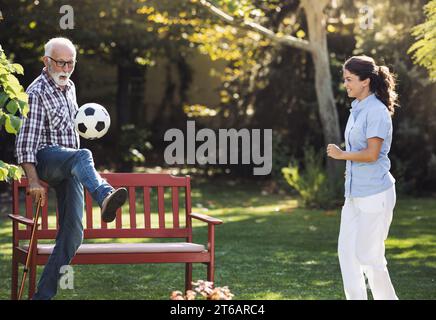 Seniorenmann mit Gehstock und junge Pflegerin, die im Park Fußball spielt Stockfoto