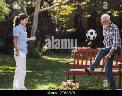 Seniorenmann mit Gehstock und junge Pflegerin, die im Park Fußball spielt Stockfoto
