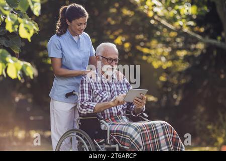 Älterer Mann im Rollstuhl, der etwas auf dem Tablet der jungen Pflegekraft hinter ihm im Park zeigt Stockfoto