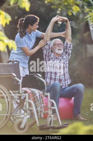 Eine junge Physiotherapeutin hilft älteren Männern beim Dehnen schmerzhafter Arme, indem sie Übungen auf dem Ball im Park, neben dem Rollstuhl, macht Stockfoto