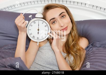 Das hübsche Mädchen hält eine Uhr und liegt im Schlafzimmer. Emotional schöne Frau, die an ihrem freien Tag im Bett liegt. Stockfoto