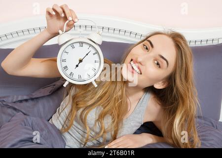 Das süße Mädchen hält eine Uhr und liegt im Schlafzimmer. Emotional schöne Frau, die an ihrem freien Tag im Bett liegt. Stockfoto