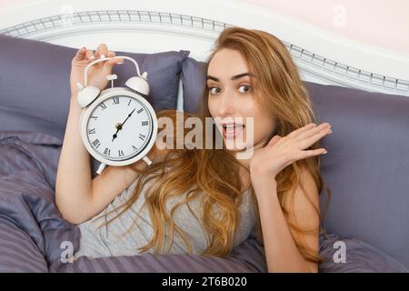 Das schöne Mädchen hält eine Uhr und liegt im Schlafzimmer. Emotional schöne Frau, die an ihrem freien Tag im Bett liegt. Stockfoto