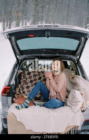 Die junge Frau sitzt mit einer Tasse Kaffee, die vor Lust im Kofferraum eines Autos in einem Winterwald schwingt Stockfoto