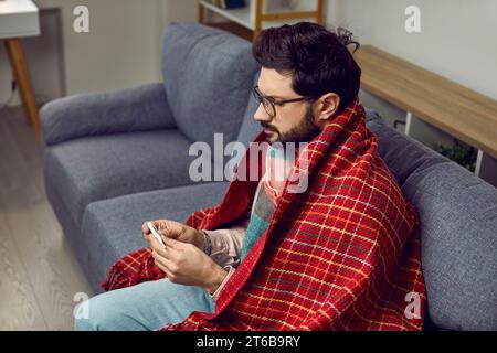 Ein kranker Mann sitzt auf der Couch, in eine Decke gewickelt, mit einem Thermometer in den Händen Stockfoto