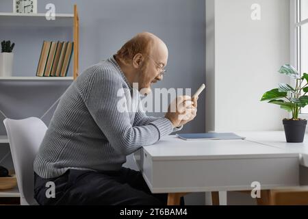 Senior-Mann sitzt zu Hause am Schreibtisch und benutzt Smartphone. Stockfoto