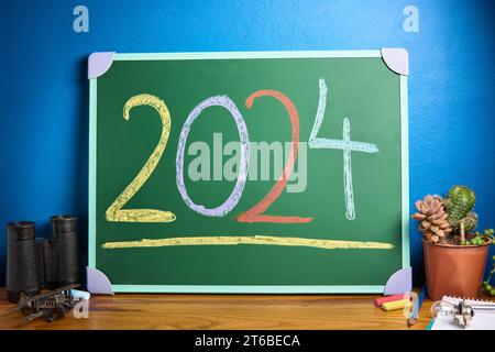 Vorderansicht der Nummer 2024, geschrieben in Kreide auf grüner Tafel auf Holztisch mit Zubehör gegen flache blaue Wand. Neujahr, zurück zu s Stockfoto