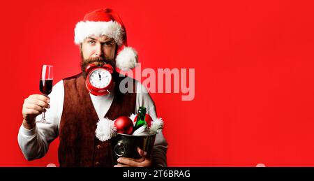 Zeit zum Feiern. Bärtiger Mann mit Weihnachtsmütze mit Wecker. Der Kellner hält ein Glas Wein und einen Eimer mit einer Flasche Champagner und Weihnachtsdekoration Stockfoto