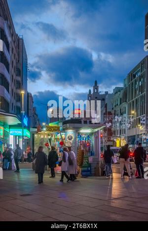 Menschen, die in der Nähe eines Kiosks für alkoholfreie Getränke und Süßigkeiten vorbeifahren, mit dunkelgrauen Wolken im Hintergrund in Callao bei Nacht Madrid Spanien Stockfoto