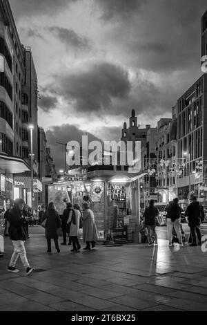 Schwarz-weiß-Stil von Menschen, die in der Nähe eines Kiosks für alkoholfreie Getränke und Süßigkeiten vorbeifahren, mit dunkelgrauen Wolken im Hintergrund in Callao bei Nacht Madrid Spanien Stockfoto