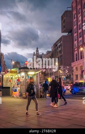 Menschen, die in der Nähe eines Kiosks für alkoholfreie Getränke und Süßigkeiten vorbeifahren, mit dunkelgrauen Wolken im Hintergrund in Callao bei Nacht Madrid Spanien Stockfoto