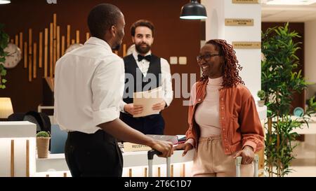 afroamerikanisches Paar, das nach einem erfolgreichen Flitterwochenurlaub aus dem Luxushotel auscheckte. Glückliche Gäste, unterstützt von einem freundlichen, hilfsbereiten Rezeptionisten an der Rezeption in der Hotellobby Stockfoto
