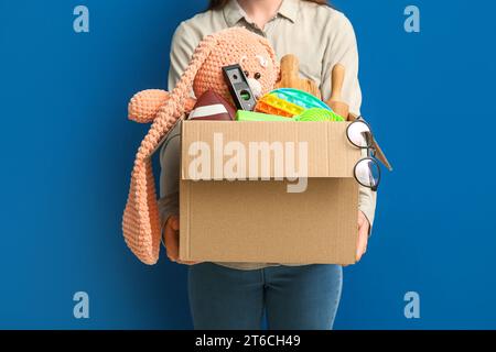 Frau, die eine Schachtel mit unerwünschten Sachen für den Hofverkauf auf blauem Hintergrund hält Stockfoto