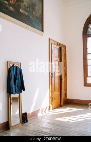 Bräutigam-Anzug hängt an einem Kleiderbügel am Spiegel neben seinen Schuhen in der großen Halle der Villa Stockfoto