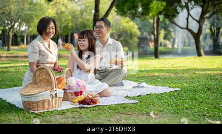 Nette und liebenswerte asiatische Großeltern genießen ein Picknick im Garten mit ihrer entzückenden kleinen Enkelin. Schönes Familienkonzept Stockfoto