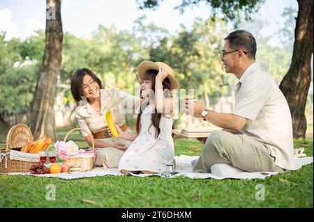 Nette und glückliche asiatische Großeltern genießen ein Picknick mit ihrer niedlichen Enkelin in einem öffentlichen Park an einem hellen Tag, sitzen auf einer Picknickdecke und Stockfoto