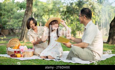 Nette und glückliche asiatische Großeltern genießen ein Picknick mit ihrer niedlichen Enkelin in einem öffentlichen Park an einem hellen Tag, sitzen auf einer Picknickdecke und Stockfoto