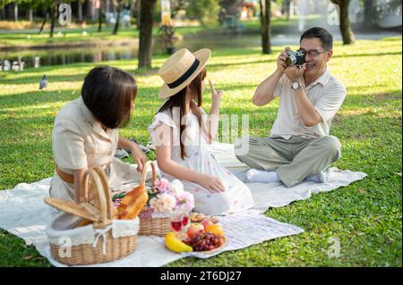 Ein glücklicher asiatischer Großvater fotografiert seine Frau und Enkelin mit seiner analogen Filmkamera, während er ein Picknick im grünen Park macht Stockfoto