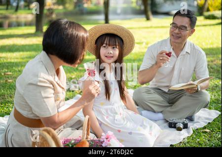 Ein entzückendes junges asiatisches Mädchen redet gerne mit ihrer Großmutter und ihrem Opa, während sie gemeinsam in einem grünen Park picknickt. Familienverbund-Konzept, großartig Stockfoto