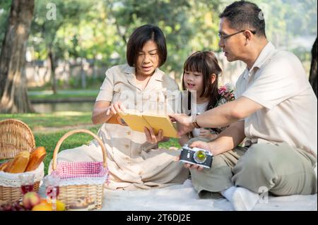 Eine glückliche und gütige asiatische Großeltern erzählen ihrer hübschen kleinen Enkelin ein Märchen in einem Buch, während sie ein Picknick in einem grünen Park genießen Stockfoto