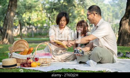 Eine glückliche und gütige asiatische Großeltern erzählen ihrer hübschen kleinen Enkelin ein Märchen in einem Buch, während sie ein Picknick in einem grünen Park genießen Stockfoto