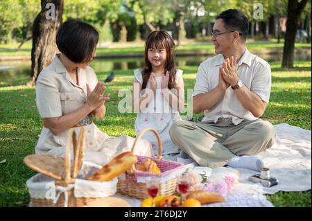 Glückliche asiatische Großeltern und Enkelinnen singen am Wochenende ein Lied, picknicken und haben gemeinsam Spaß im grünen öffentlichen Park. Familienleis Stockfoto