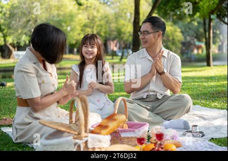 Glückliche asiatische Großeltern und Enkelinnen singen am Wochenende ein Lied, picknicken und haben gemeinsam Spaß im grünen öffentlichen Park. Familienleis Stockfoto