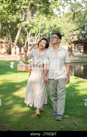 Schöne und glückliche asiatische Seniorenpaare halten Hände und genießen es, an einem hellen Tag gemeinsam durch den grünen Park zu schlendern. Glückliches Heiratsleben Stockfoto