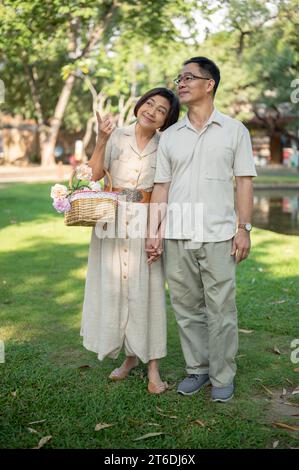 Schöne und glückliche asiatische Seniorenpaare halten Hände und genießen es, an einem hellen Tag gemeinsam durch den grünen Park zu schlendern. Glückliches Heiratsleben Stockfoto