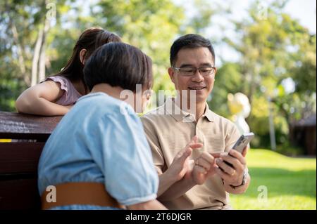 Liebevolle und freundliche ältere asiatische Großeltern und süße kleine Enkelin benutzen gemeinsam ein Smartphone, sitzen auf einer Bank und entspannen sich in einem Park Stockfoto