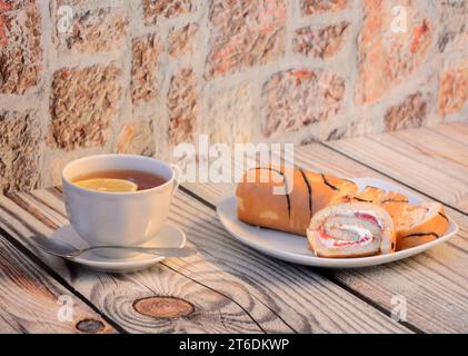 Ein Teller mit Stücken Keksrolle mit Proteincreme und einer Tasse heißen Tee mit Zitrone auf einem Holztisch. Nahaufnahme. Stockfoto