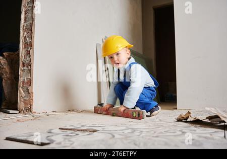 Niedlicher Bauarbeiter, der sich in einem Raum mit weißen Wänden hockt und das Wasserwaage hält. Kind trägt Schutzhelm und Arbeitsanzug während der Renovierung. Stockfoto